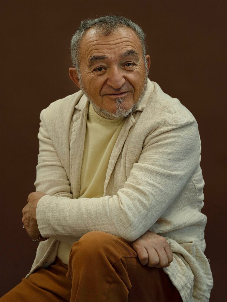 Portrait of a smiling older man with a beard, wearing a light-colored jacket and sweater, seated against a brown background, representing artistic expression and cultural engagement.
